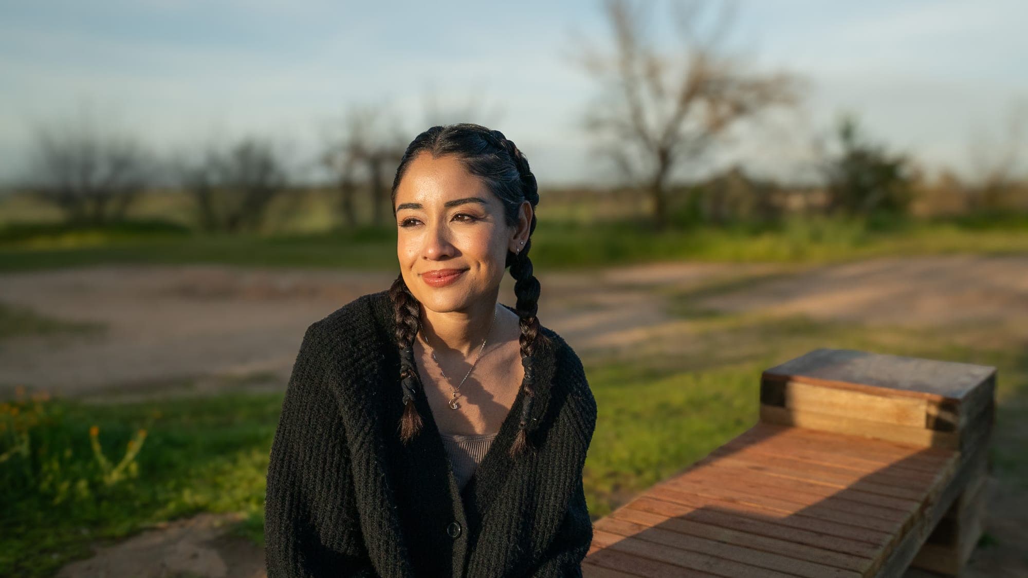 Sunlit portrait of a woman with braids sitting near a wooden bench in open countryside