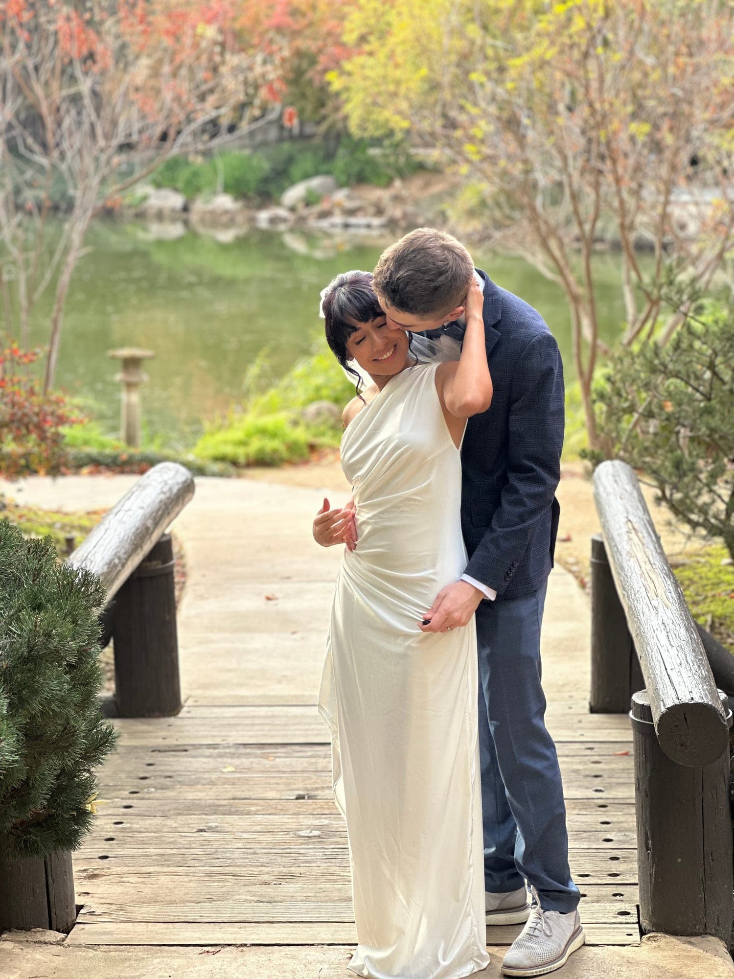 Groom embracing his bride from behind beside an autumn garden pond