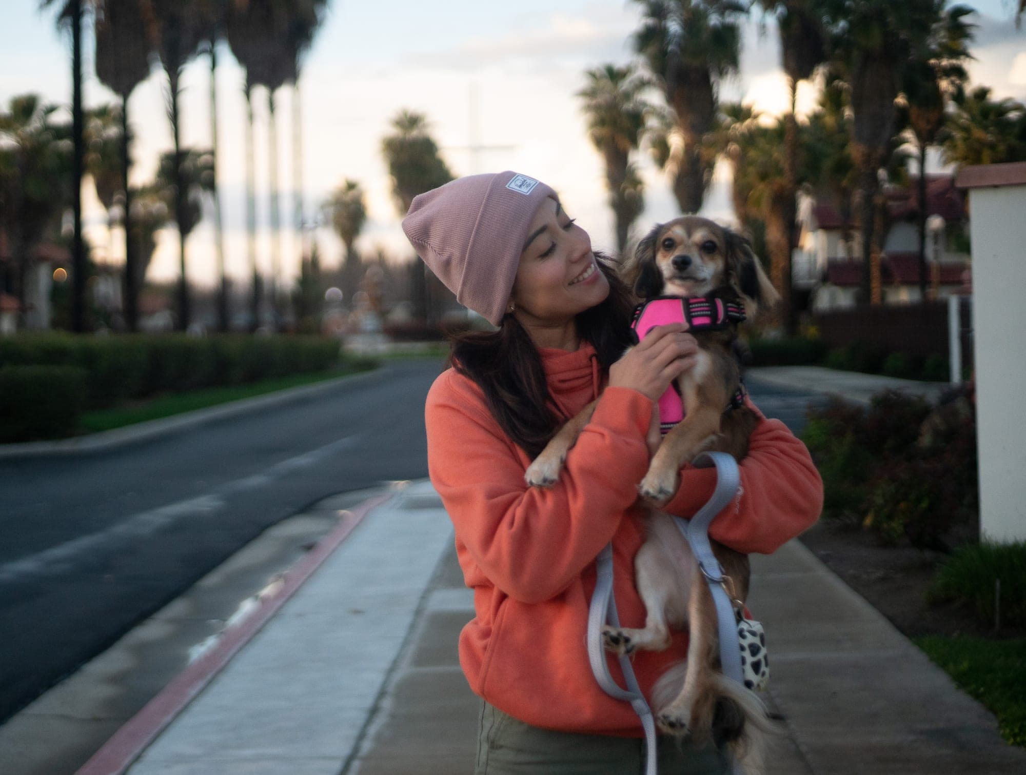 Woman in an orange hoodie laughing with her small dog under Fresno palm trees at sunset